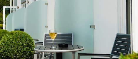 A small outdoor seating area with a table and chairs, a glass of white wine, and a neatly trimmed hedge.