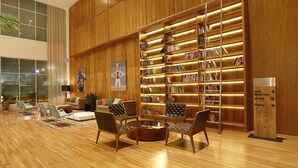 A spacious lobby with a wooden floor, a central seating area with chairs and a coffee table, a large bookshelf, and a sign indicating directions to various areas. - Vitória Hotel Convention Paulínia (Paulinia)