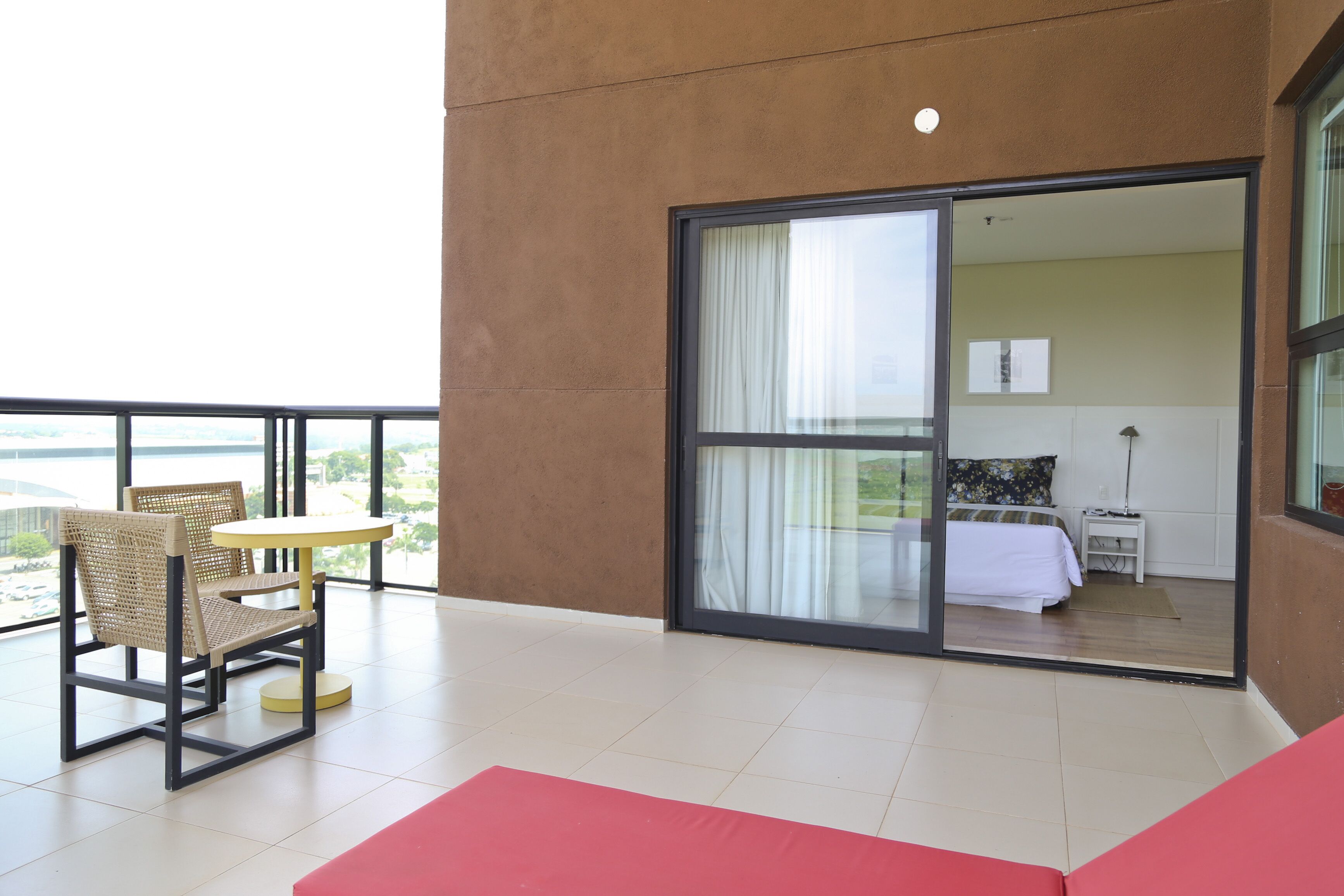 Varanda com cadeira de vime e uma pequena mesa, porta de vidro deslizante dando para um quarto com cama e mesa de cabeceira, e vista para o exterior.