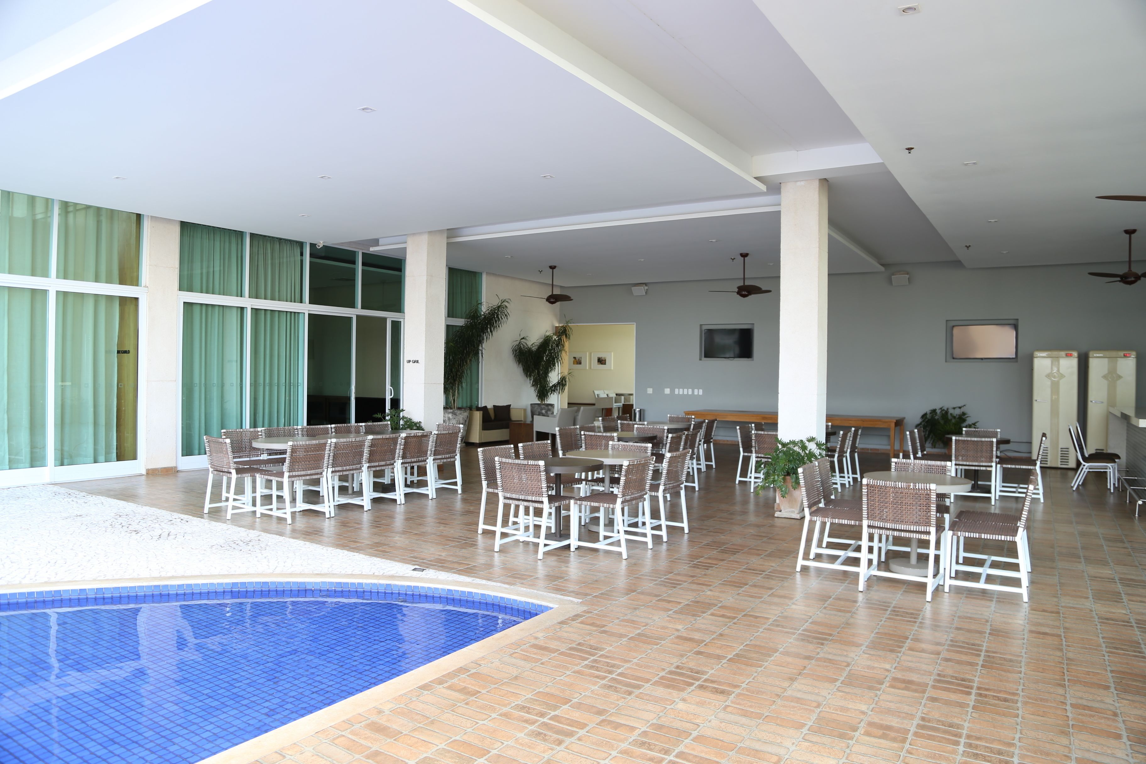 Un área de piscina interior con suelo de baldosas, varios conjuntos de asientos y ventanas amplias con cortinas verdes.