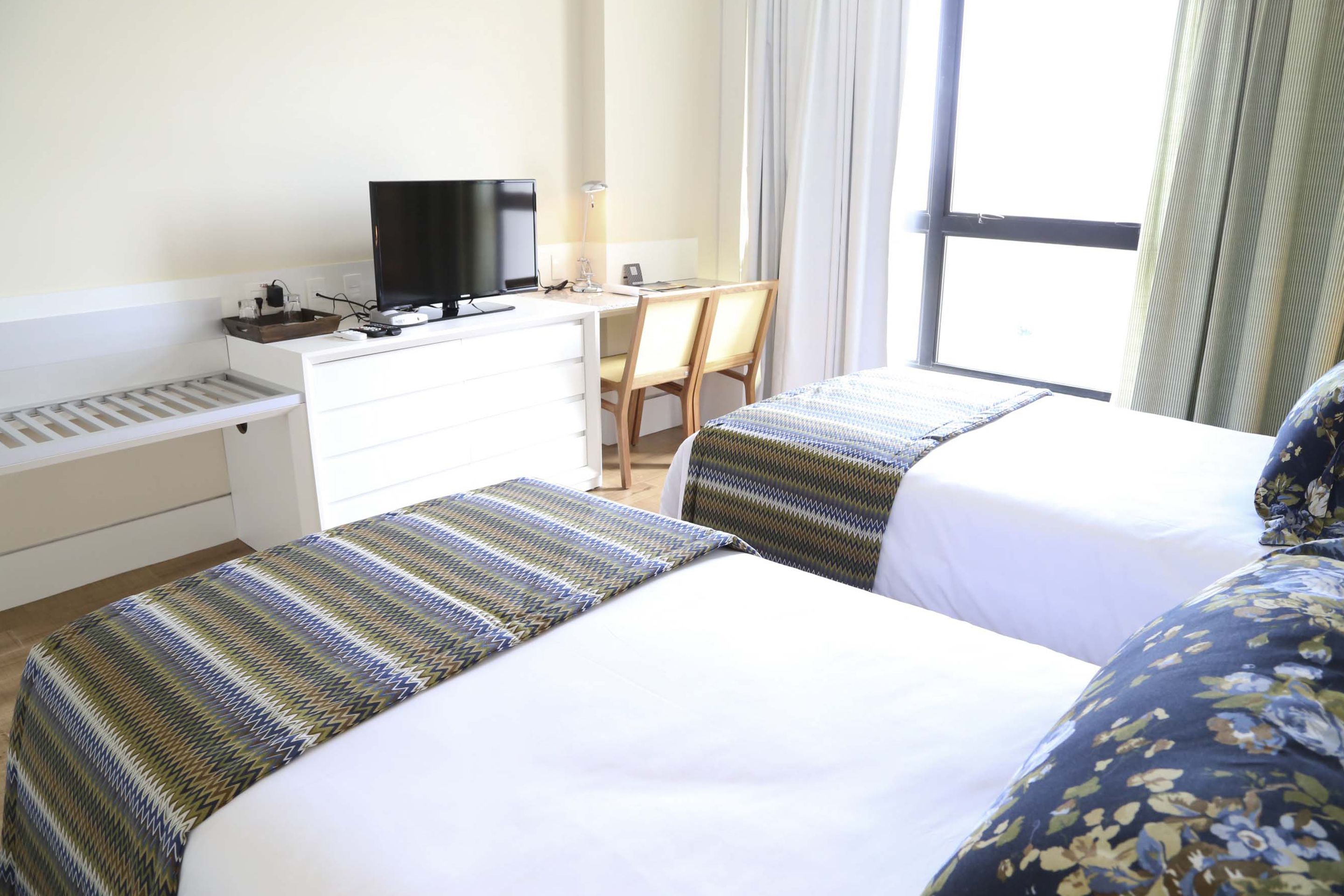 A hotel room with two beds, a desk, a television, and a window with curtains.