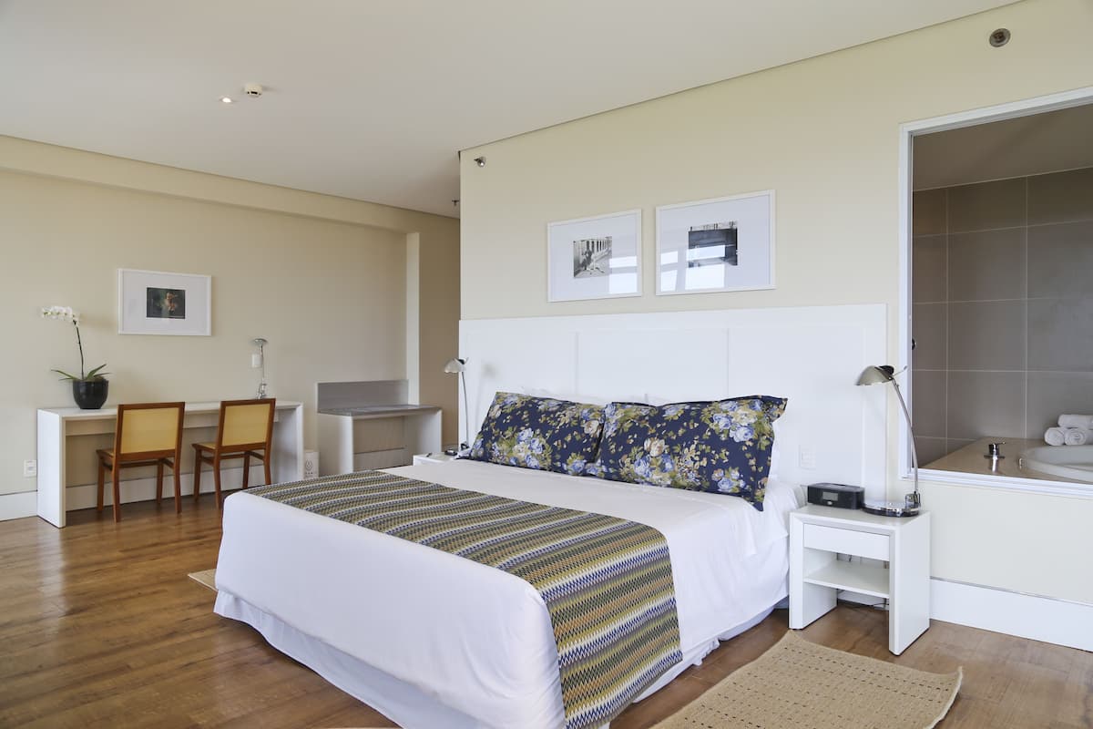 Een hotelkamer met een groot bed, een bureau, twee stoelen, een kleine tafel, een nachtkastje, een lamp en een badkamer met een bad.