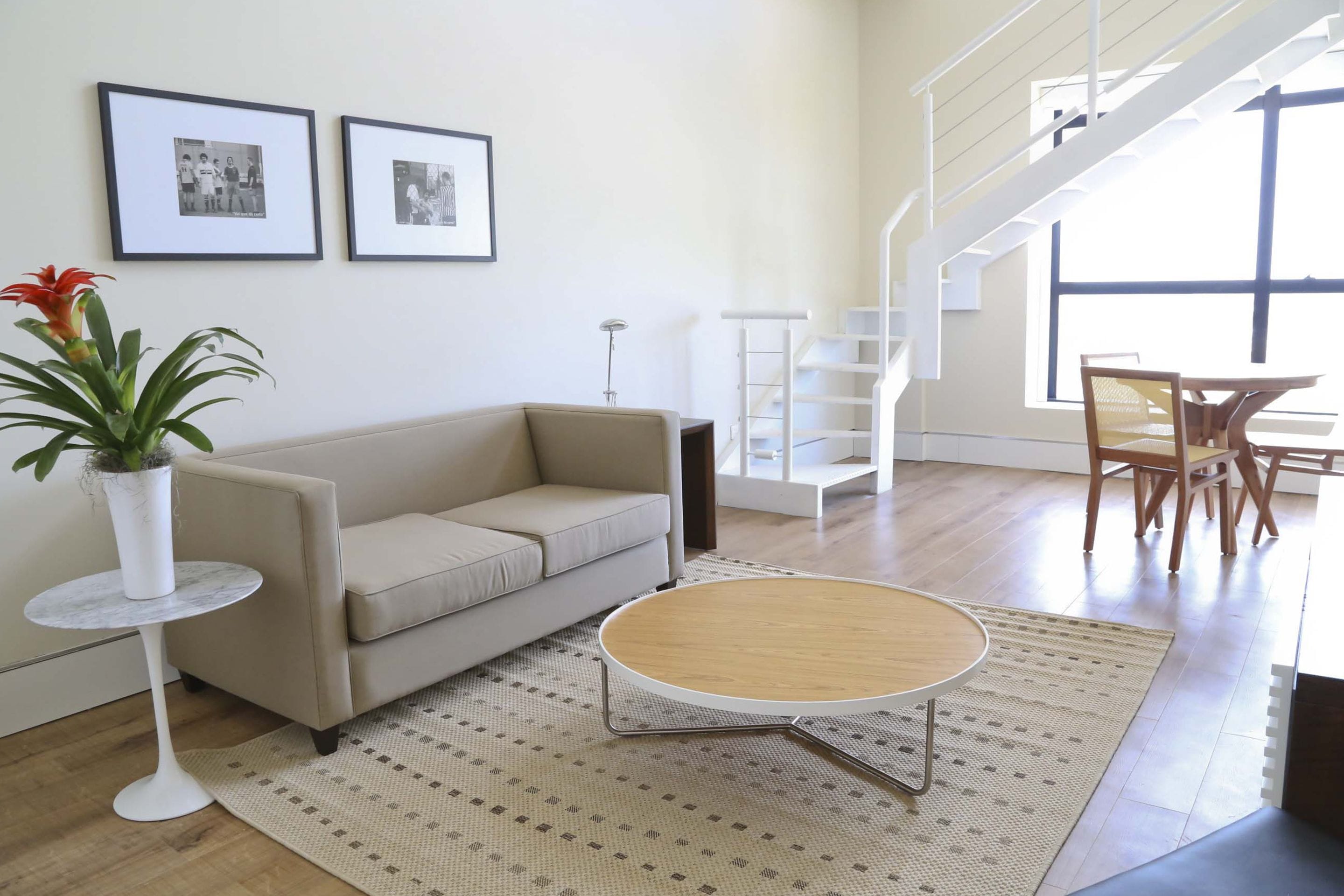 A modern living room with a beige sofa, a round wooden coffee table, a small white side table with a plant, and a patterned rug. There are two framed pictures on the wall, a white shelving unit, and a staircase leading to another level.