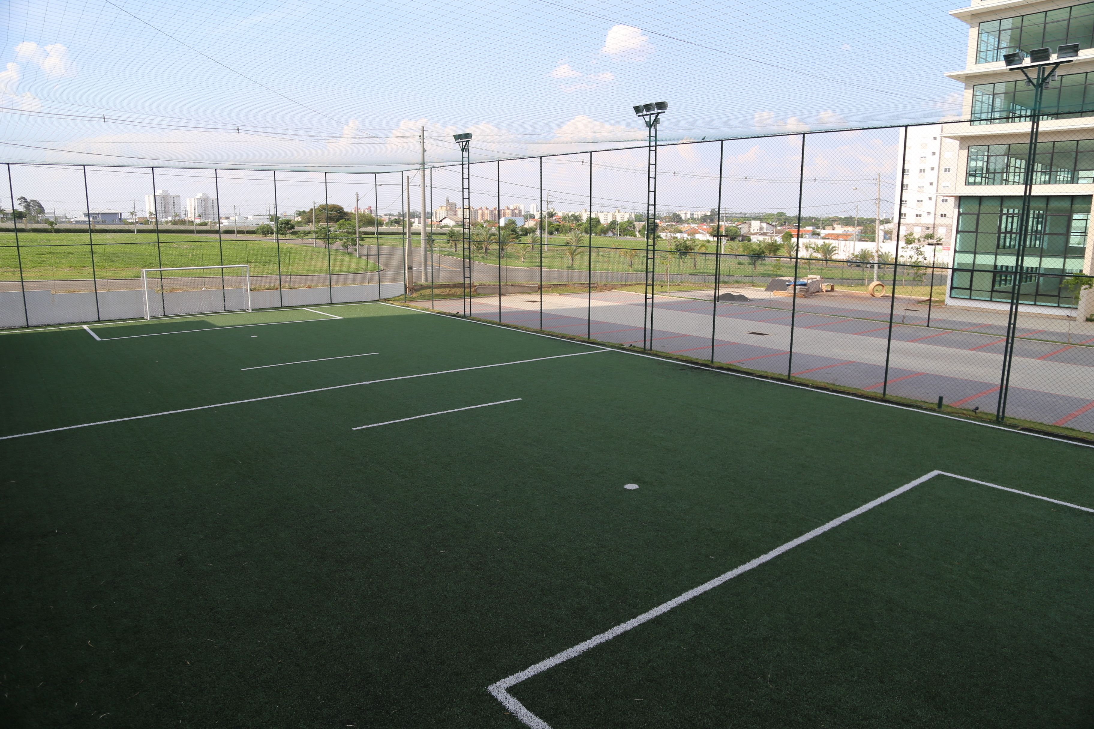 Instalación deportiva interior con césped verde, rodeada por una valla metálica y contigua a una carretera con edificios al fondo.