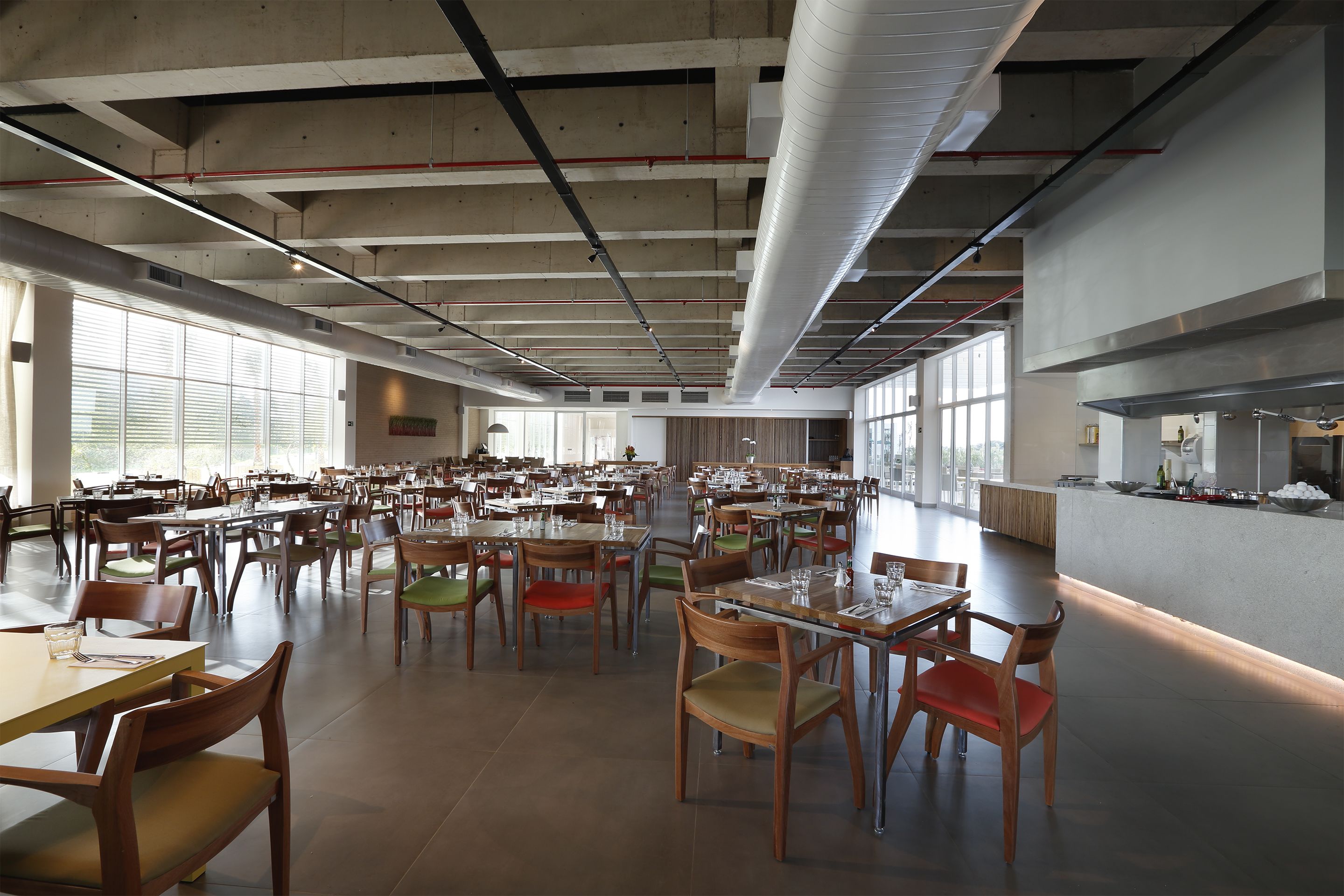 A spacious cafeteria with multiple tables and chairs, large windows, and a kitchen area.