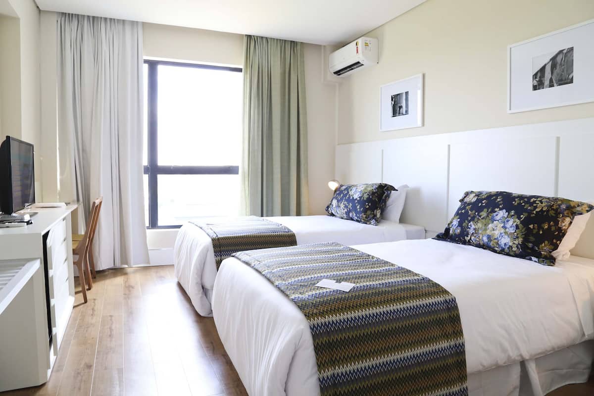 Een hotelkamer met twee bedden, een bureau en een stoel. Er is een raam met gordijnen, een airconditioning en prenten in lijsten aan de muur.