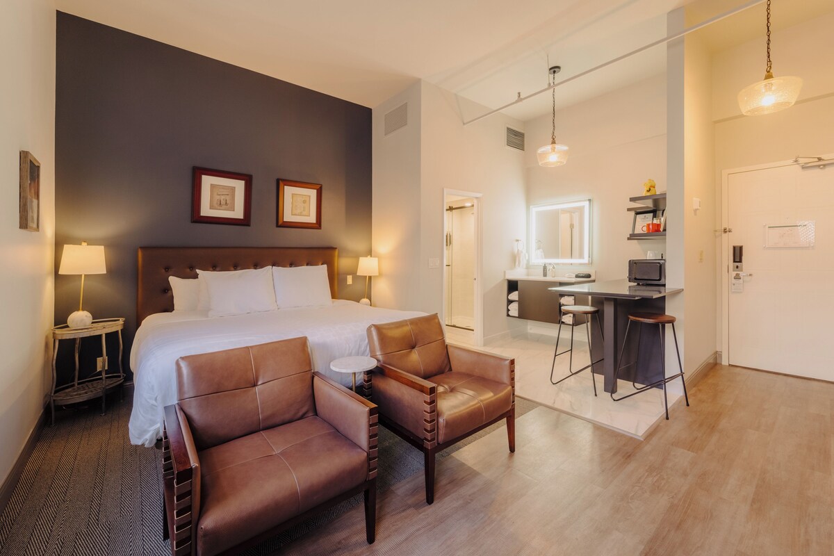 Een hotelkamer met een groot bed, twee leren fauteuils, een klein rond tafeltje, een bureau met stoel en een kitchenette.
