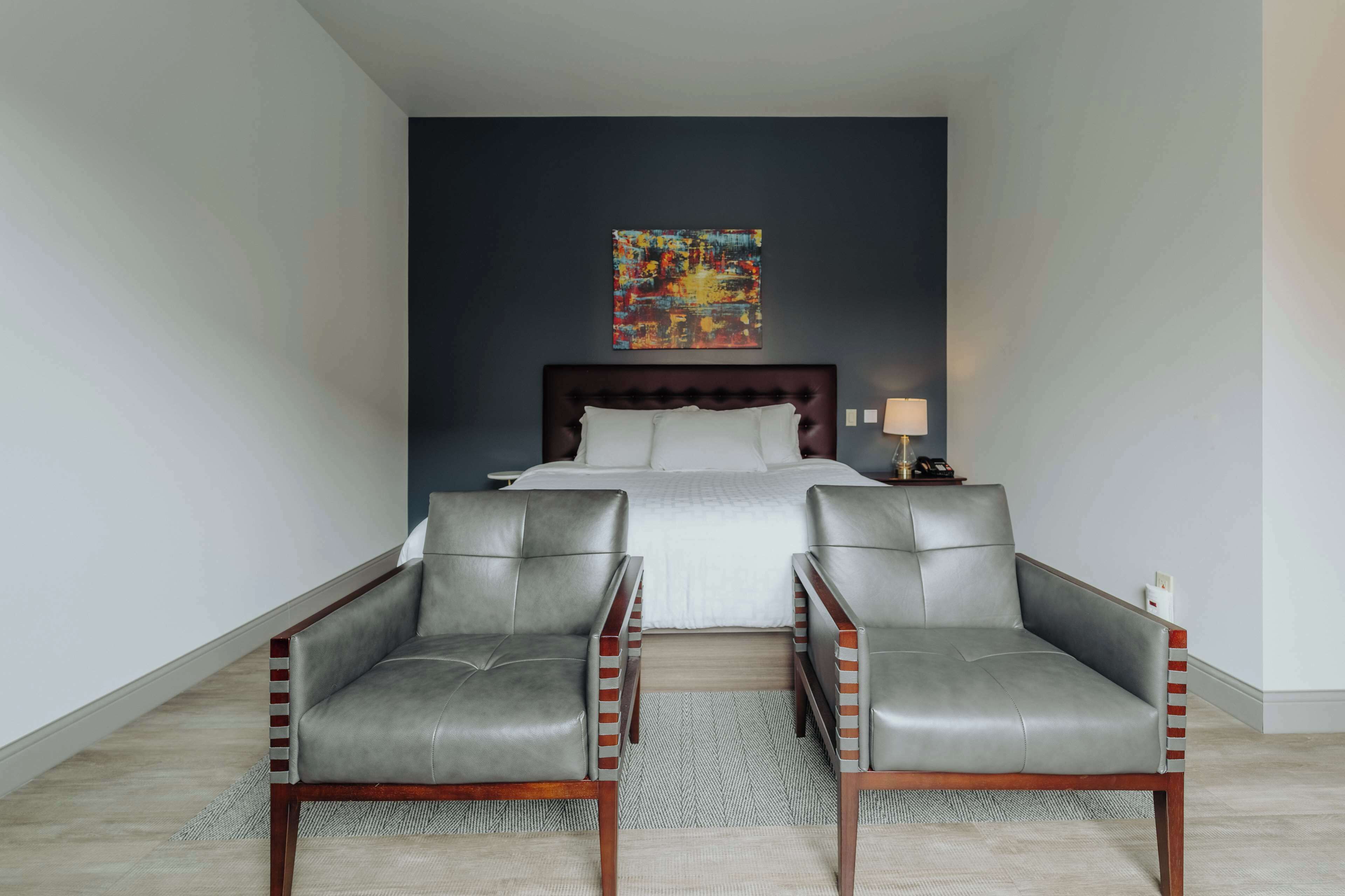 A bedroom with a bed, two grey leather chairs, a nightstand, and a colorful abstract painting on the wall.