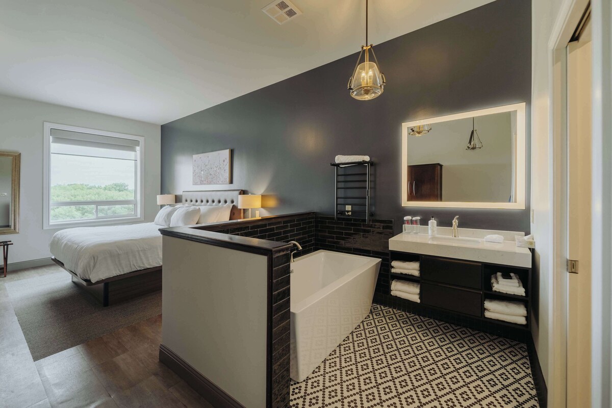 Un dormitorio moderno con una cama grande, un baño separado con bañera y un espejo amplio.
