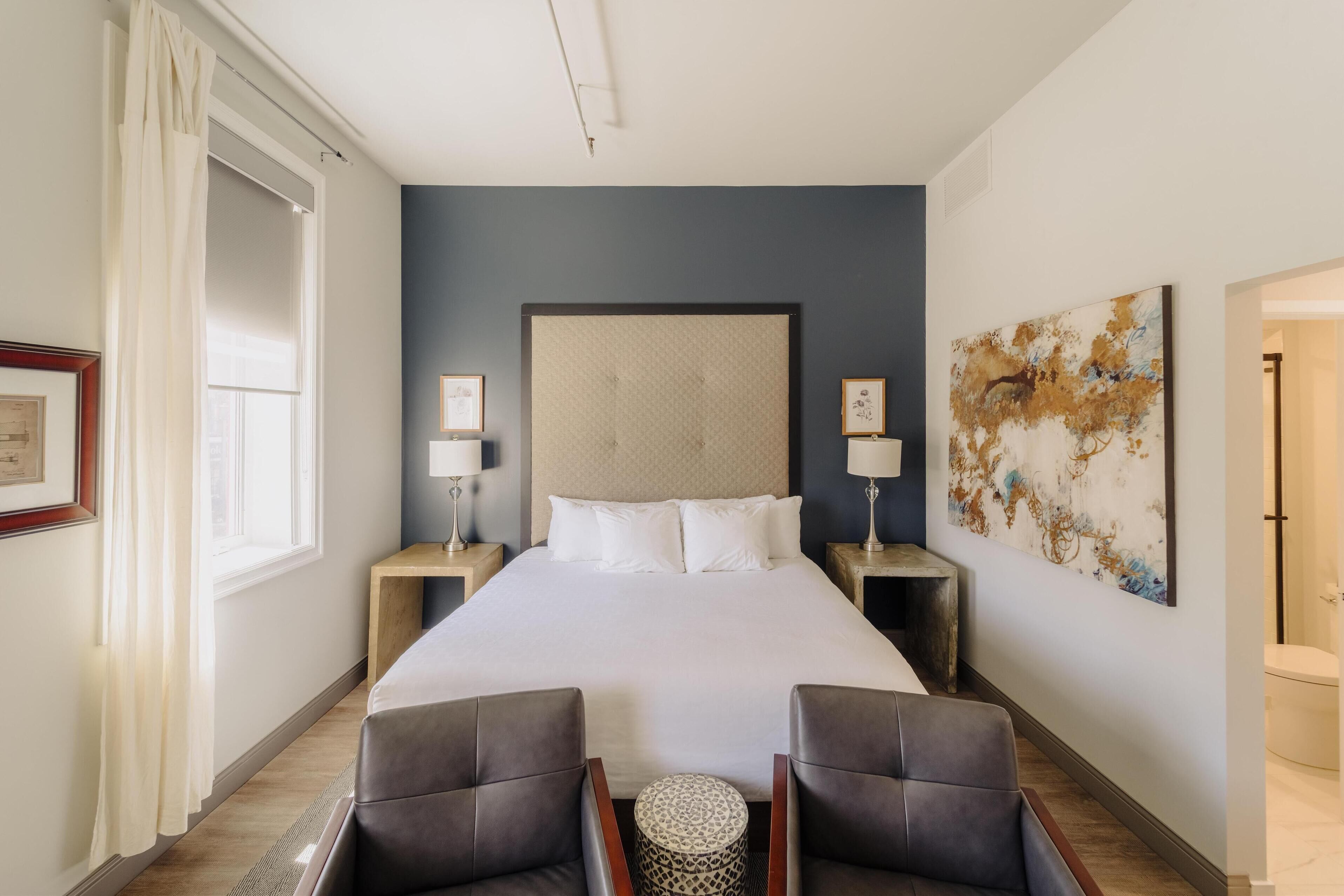 A hotel room with a large bed, two bedside tables with lamps, a headboard, a framed picture, a round side table, and a large abstract painting on the wall.