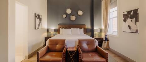 A bedroom with a large bed, two leather chairs, a small table, and a nightstand with a lamp.