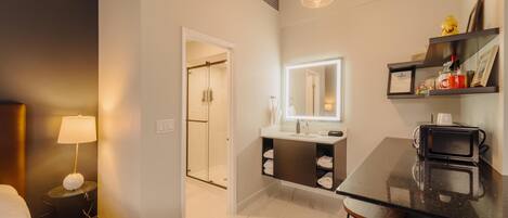 A modern hotel room with a glass door leading to a shower area, a small round table with a lamp, a bar area with stools, and a mirror above the sink.