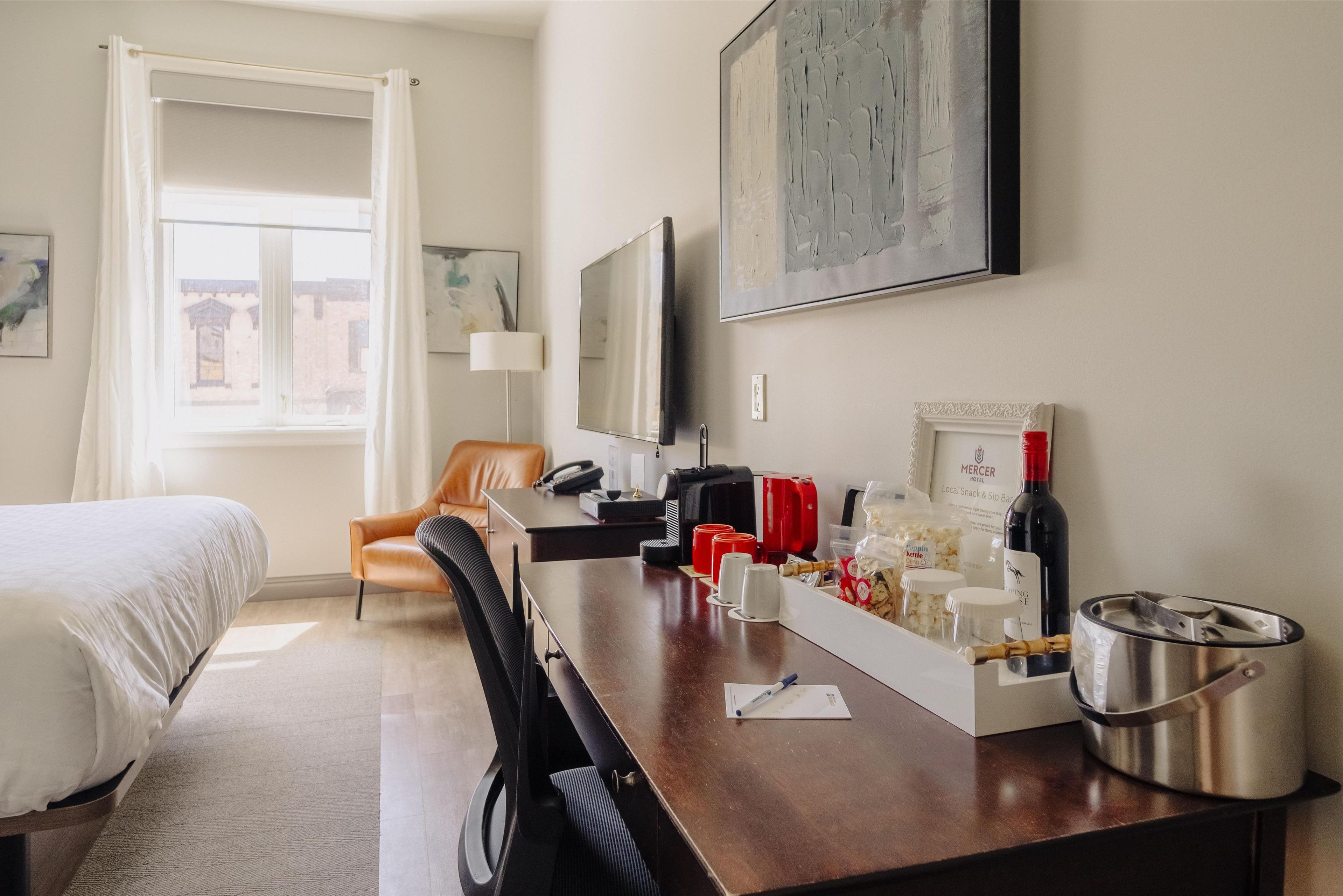 A hotel room with a bed, a desk with a chair, a television, a window with curtains, and a painting on the wall.