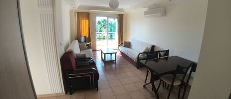 Une chambre d’hôtel avec un canapé, une table à manger, des chaises, une télévision et un balcon avec des rideaux.