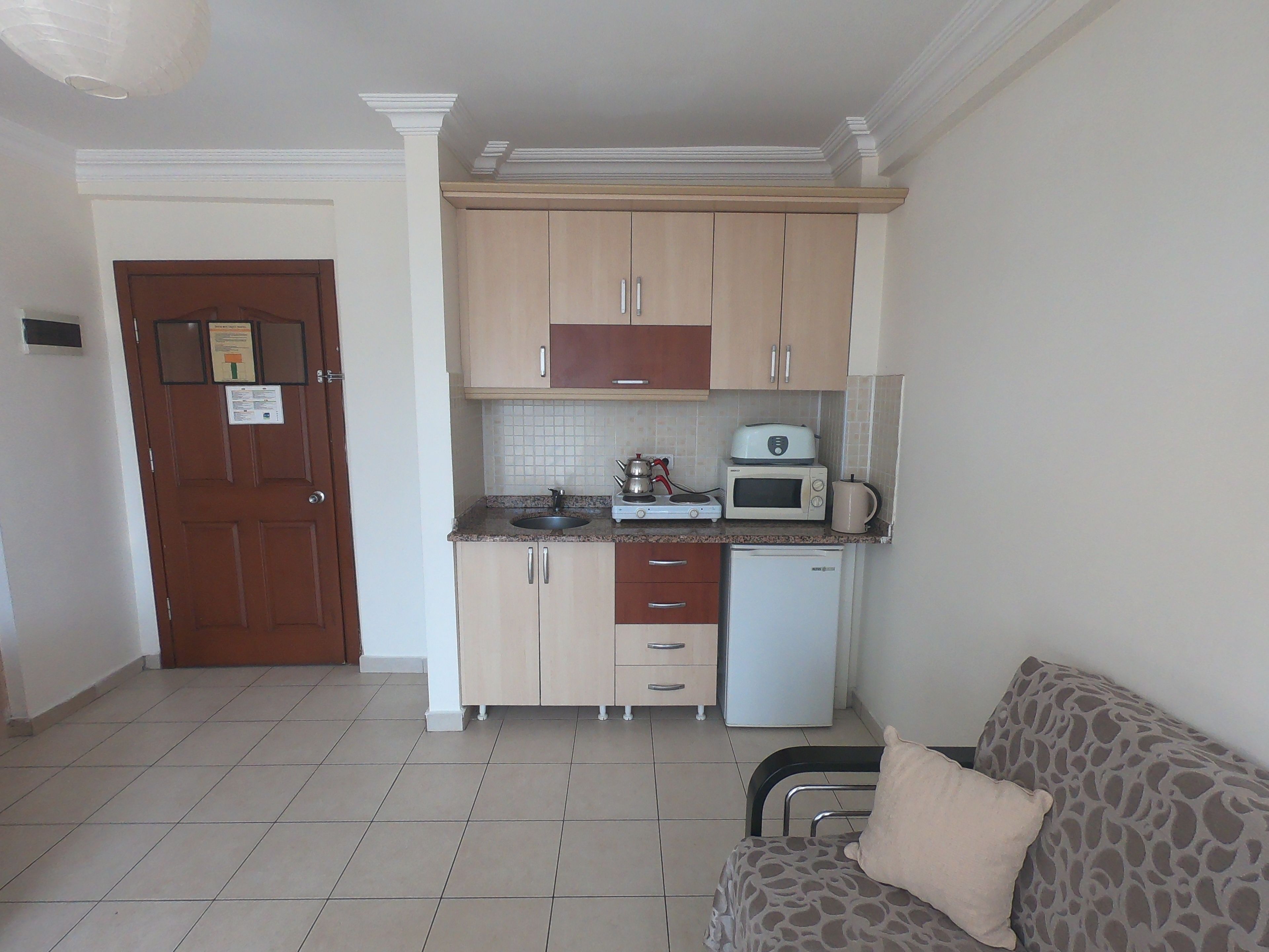 A compact kitchen with a microwave, toaster, and sink. There is a wooden door with a notice on it, and a sofa with a patterned cover in the living area.