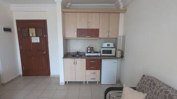 A compact kitchen with a microwave, toaster, and sink. There is a wooden door with a notice on it, and a sofa with a patterned cover in the living area.