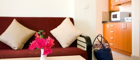 A living area with a sofa, a table with a vase of flowers, and a kitchen in the background.