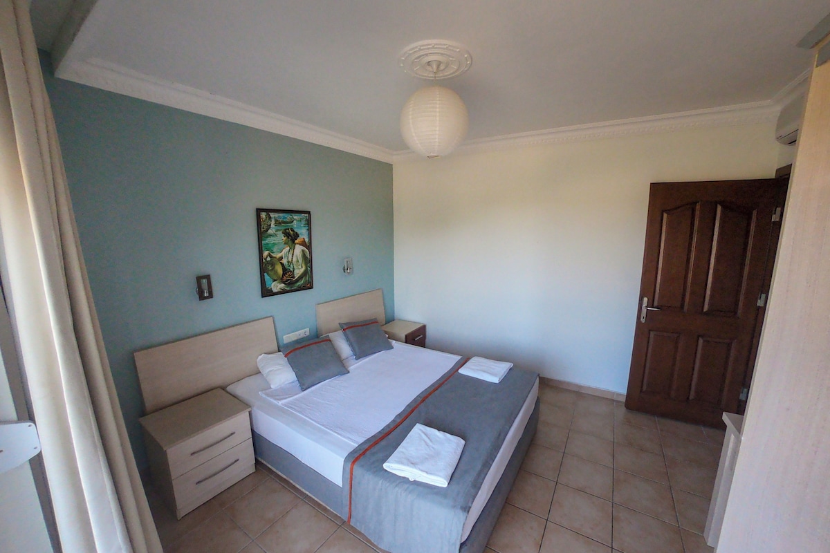 A bedroom with a large bed, two bedside tables, a nightstand, a wall-mounted light, and a door.