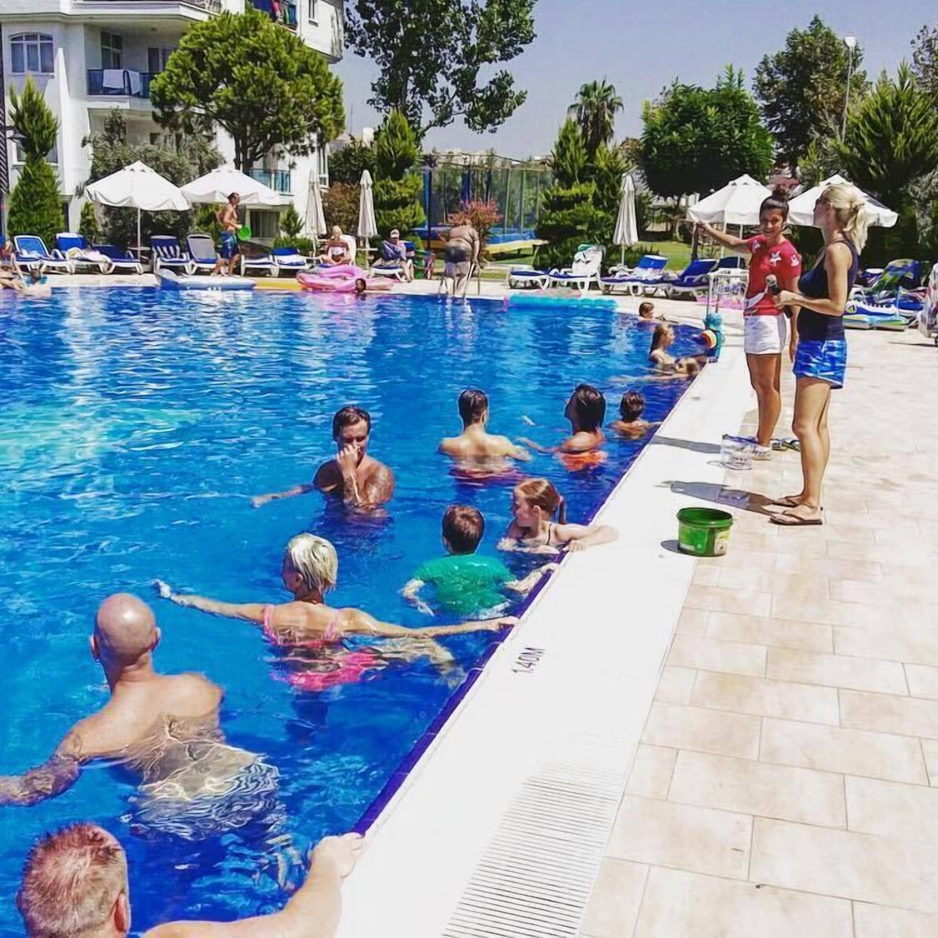 A swimming pool with people in the water and on the edge, surrounded by lounge chairs and umbrellas.