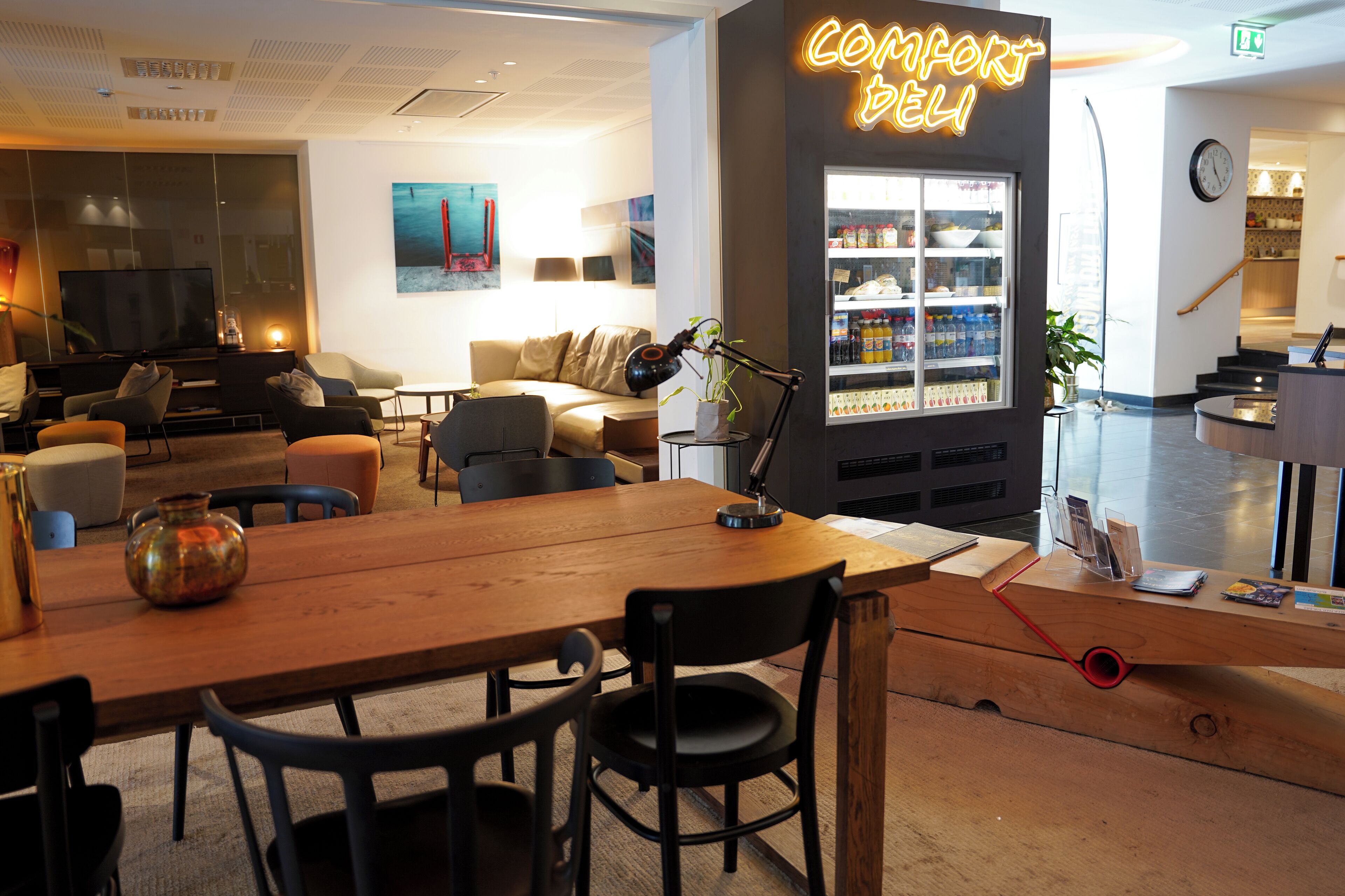 A modern hotel lobby with a wooden reception desk, a refrigerator stocked with beverages, a comfortable seating area with sofas and a TV, and a neon sign reading "COMFORT DELI".