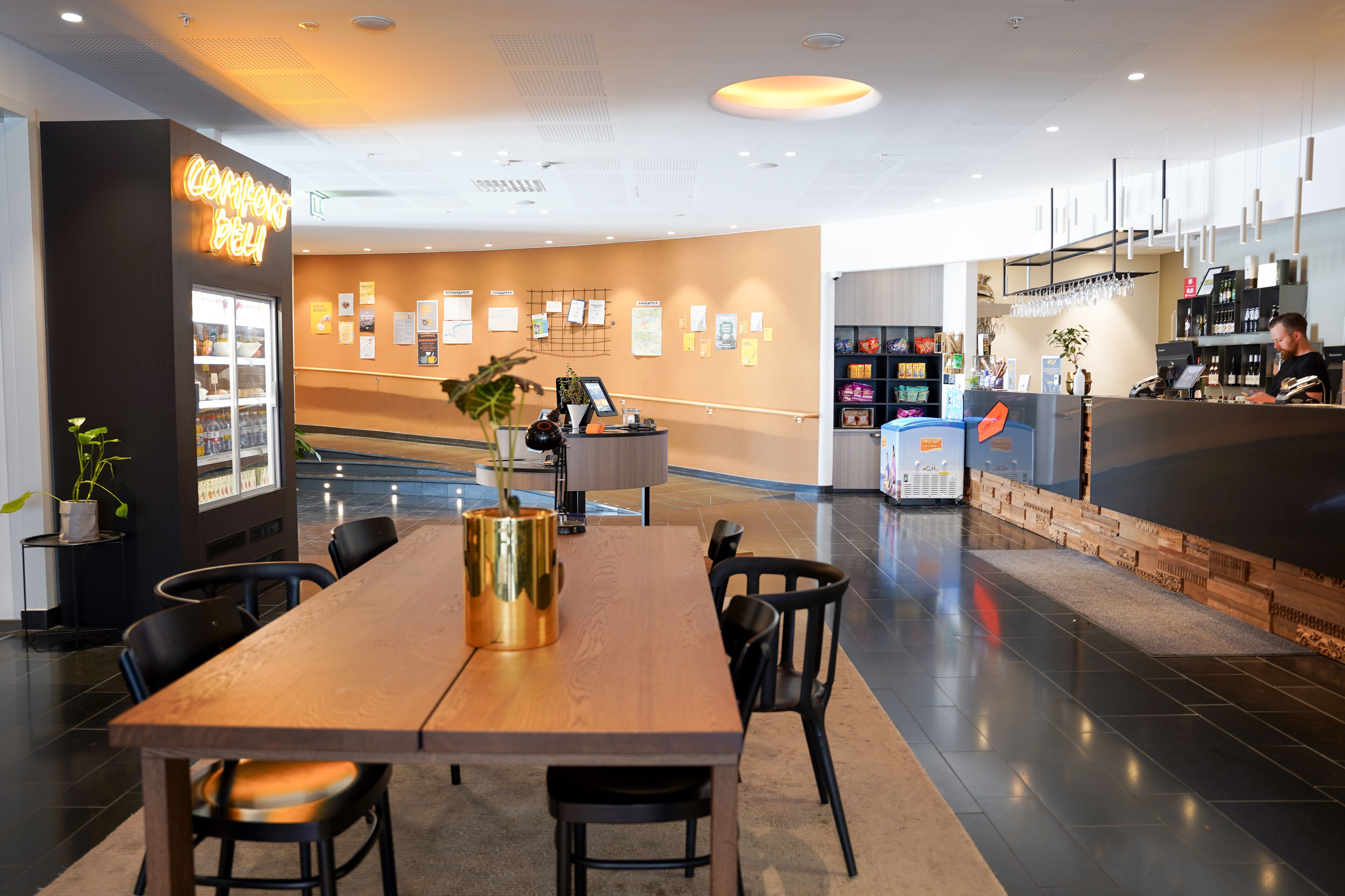 Un hall d’hôtel moderne avec un comptoir d’accueil en bois, un réfrigérateur contenant des boissons et un espace détente avec des chaises et une table.