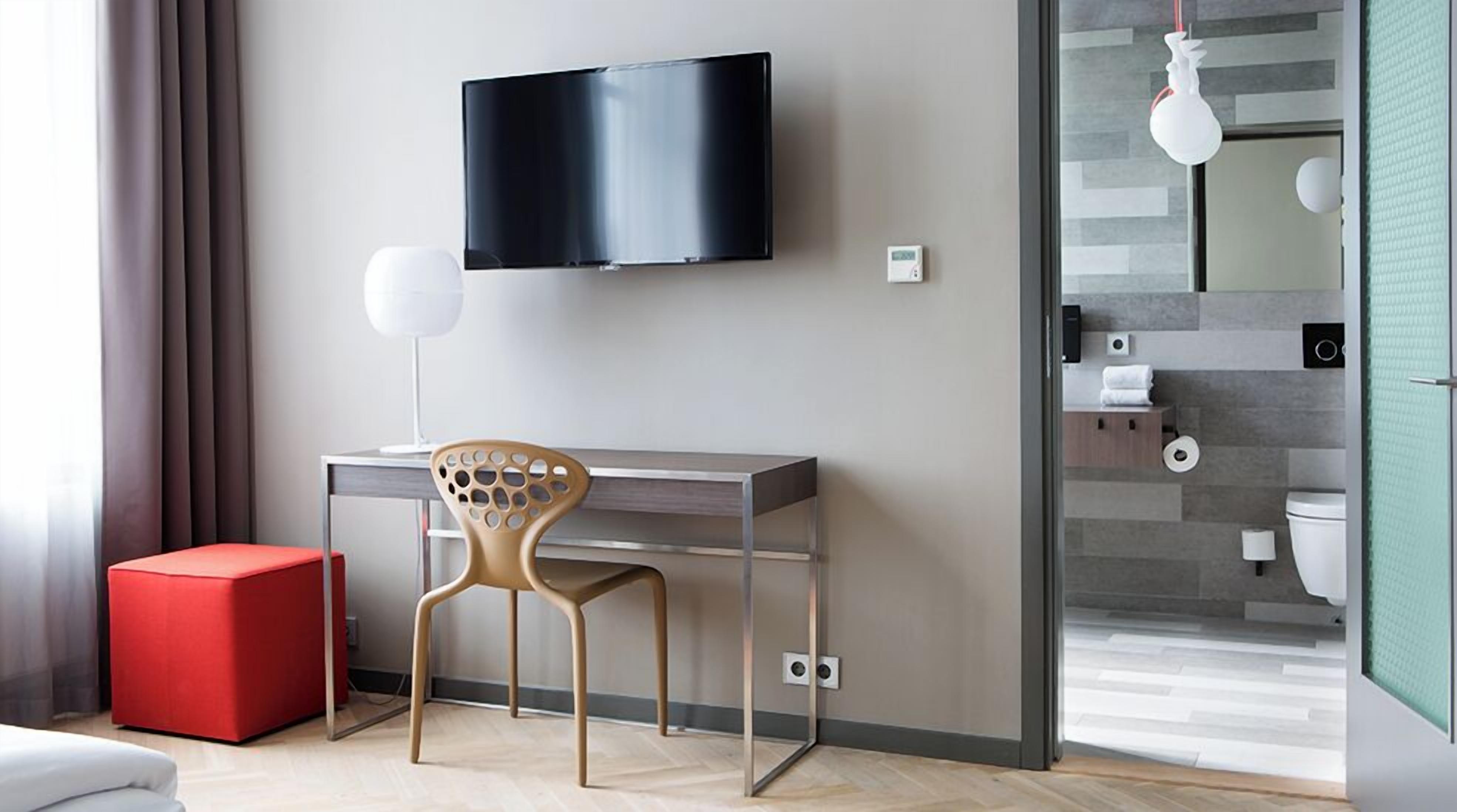 Una habitación de hotel moderna con una televisión de pantalla plana montada en la pared, un escritorio con una silla, un reposapiés rojo y un baño visible a través de un mamparo de vidrio.