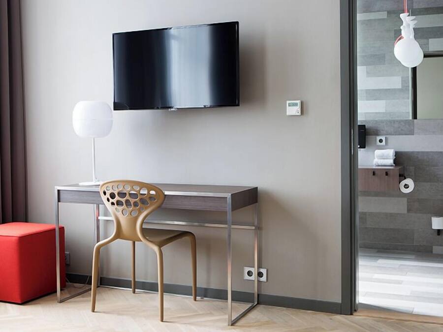 Una habitación de hotel moderna con una televisión de pantalla plana montada en la pared, un escritorio con una silla, un reposapiés rojo y un baño visible a través de un mamparo de vidrio.