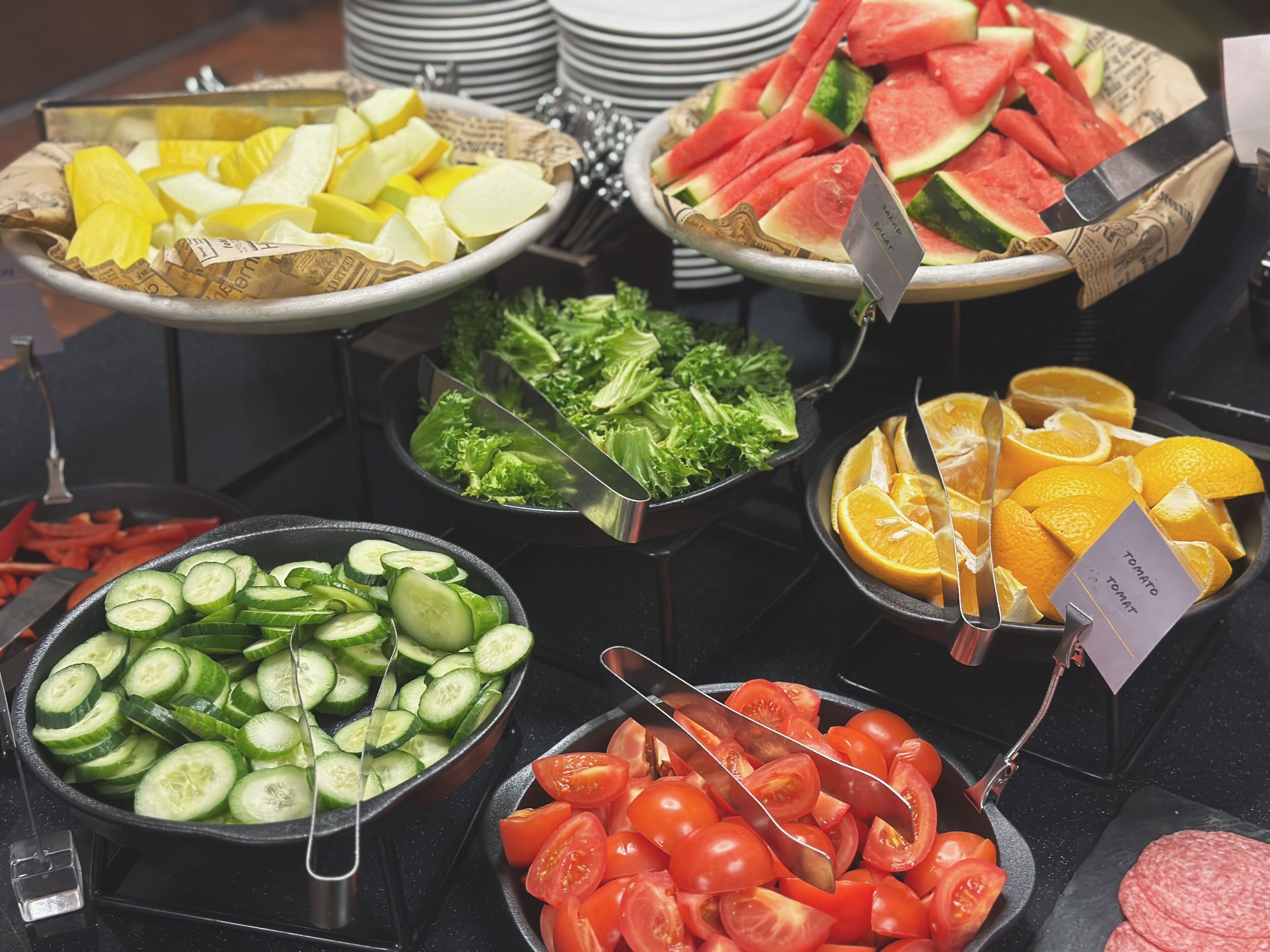 A buffet with various food items including sliced cucumbers, tomatoes, watermelon, and oranges.
