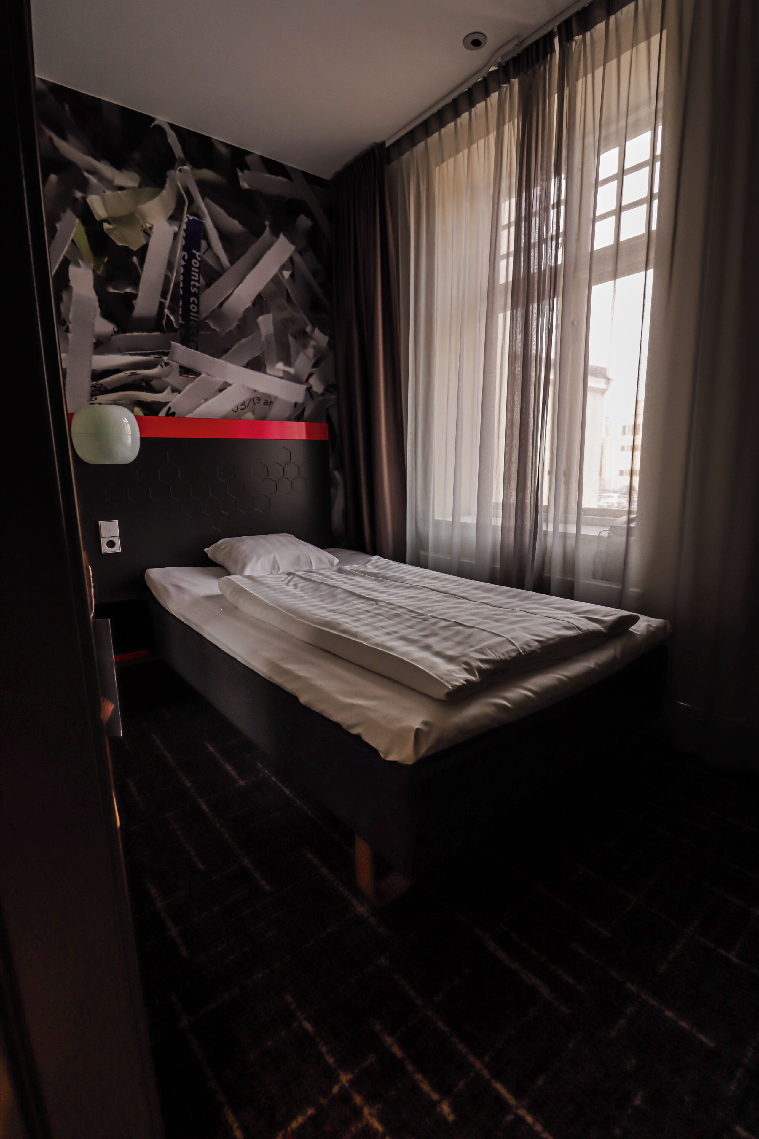 Een enkel bed met een witte matras en kussens, een muur met een honingraatpatroon, een rode plank en dunne gordijnen voor een raam.