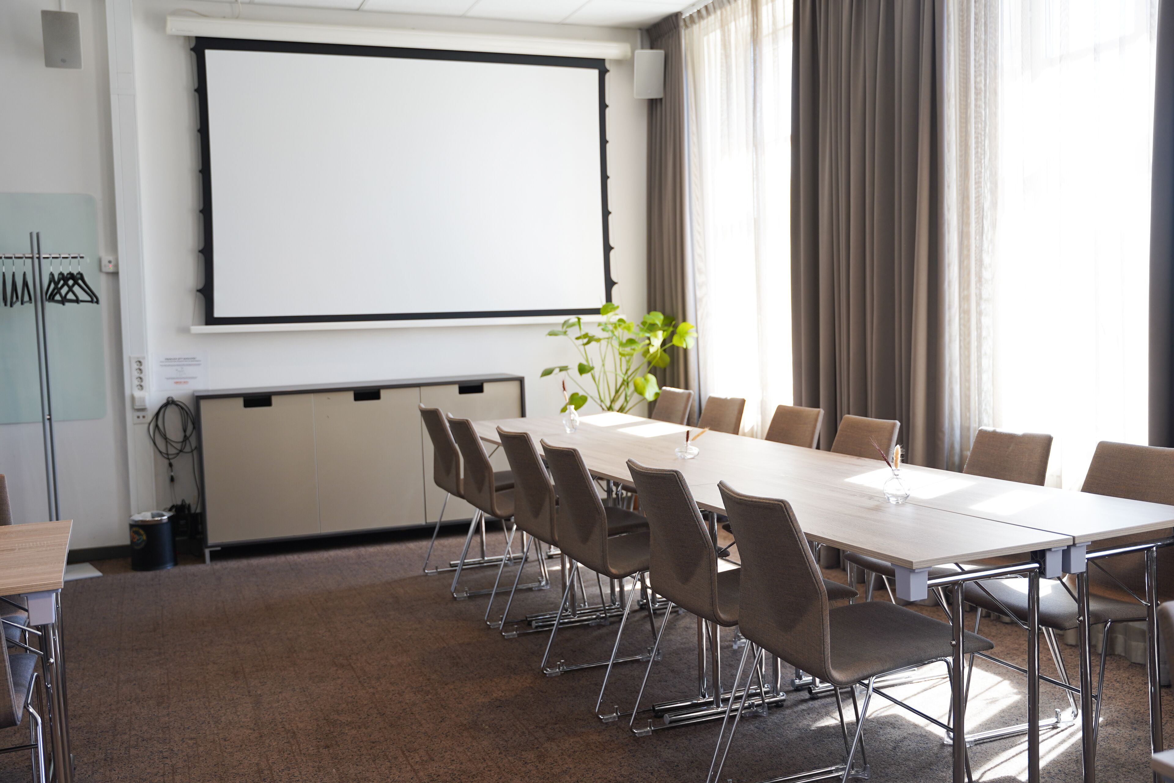 A conference room with a long table, chairs, a projector screen, and a plant.