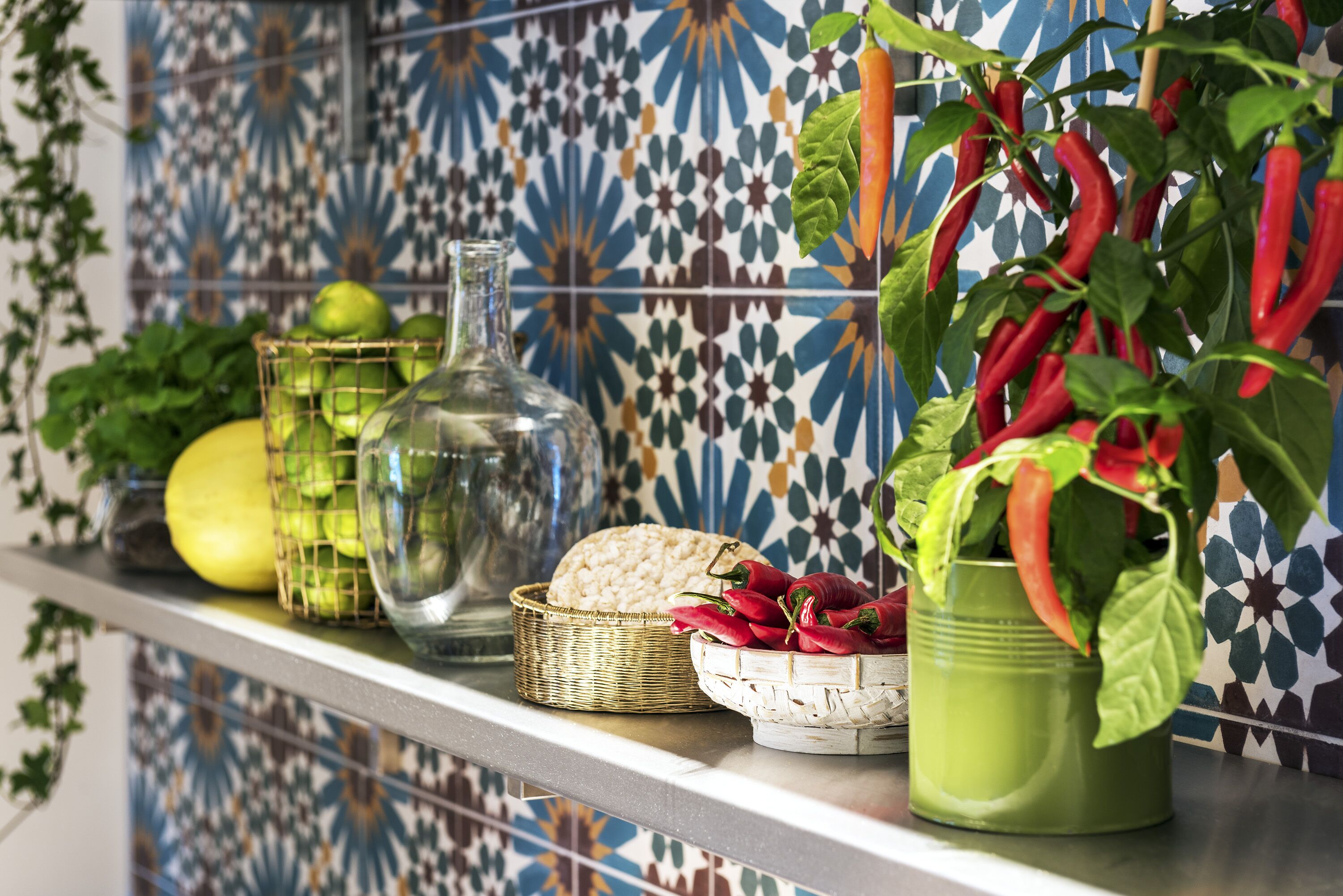 Un comptoir de cuisine agrémenté d’un pare-feu en carreaux décoratifs, une bouteille en verre, un panier en osier et une plante en pot ornée de piments rouges.