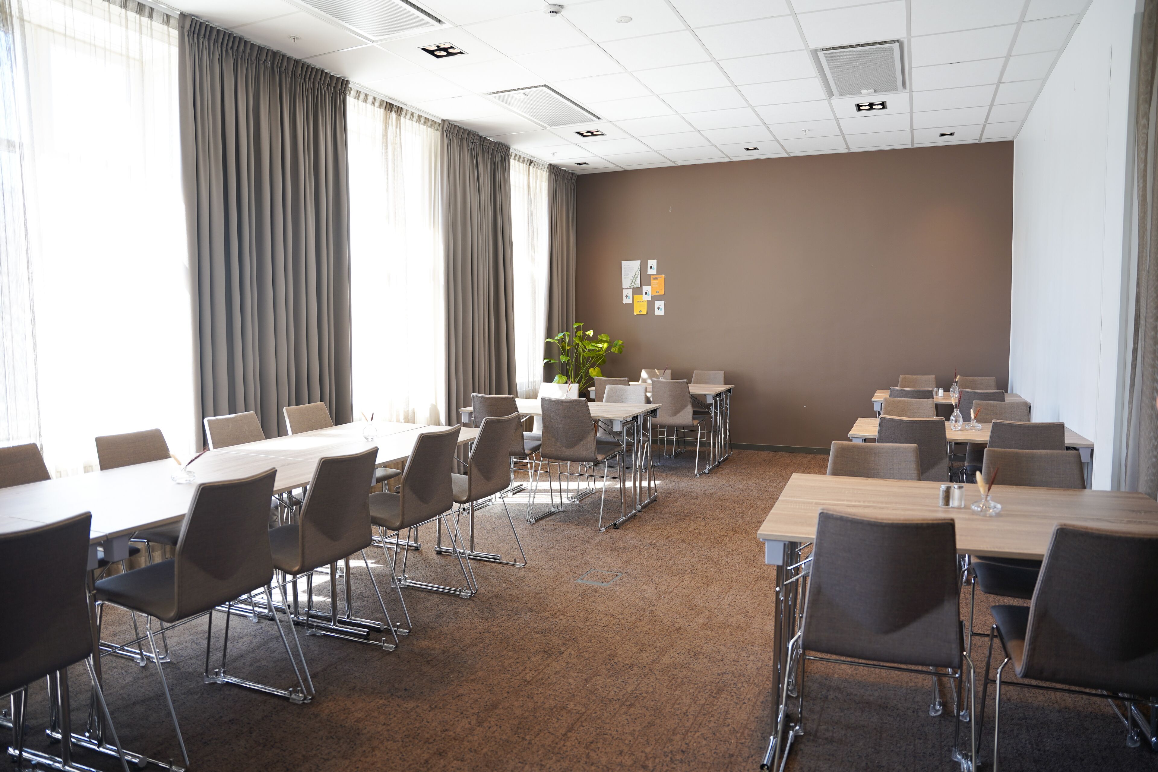 Une salle de conférence dotée de plusieurs tables et chaises, de grandes fenêtres avec des rideaux et d’un mur brun orné de quelques œuvres d’art.