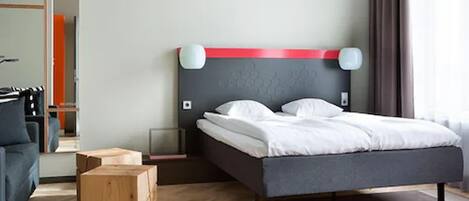 Una habitación de hotel moderna con una cama grande, un cabecero gris y un detalle rojo. Se observa un aparador de madera y un sofá gris.