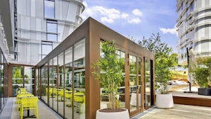 A modern outdoor seating area with yellow chairs and a glass enclosure, surrounded by potted plants and a building with a unique cylindrical design. - Ibis Styles Paris Charles de Gaulle Airport (Tremblay-en-France)