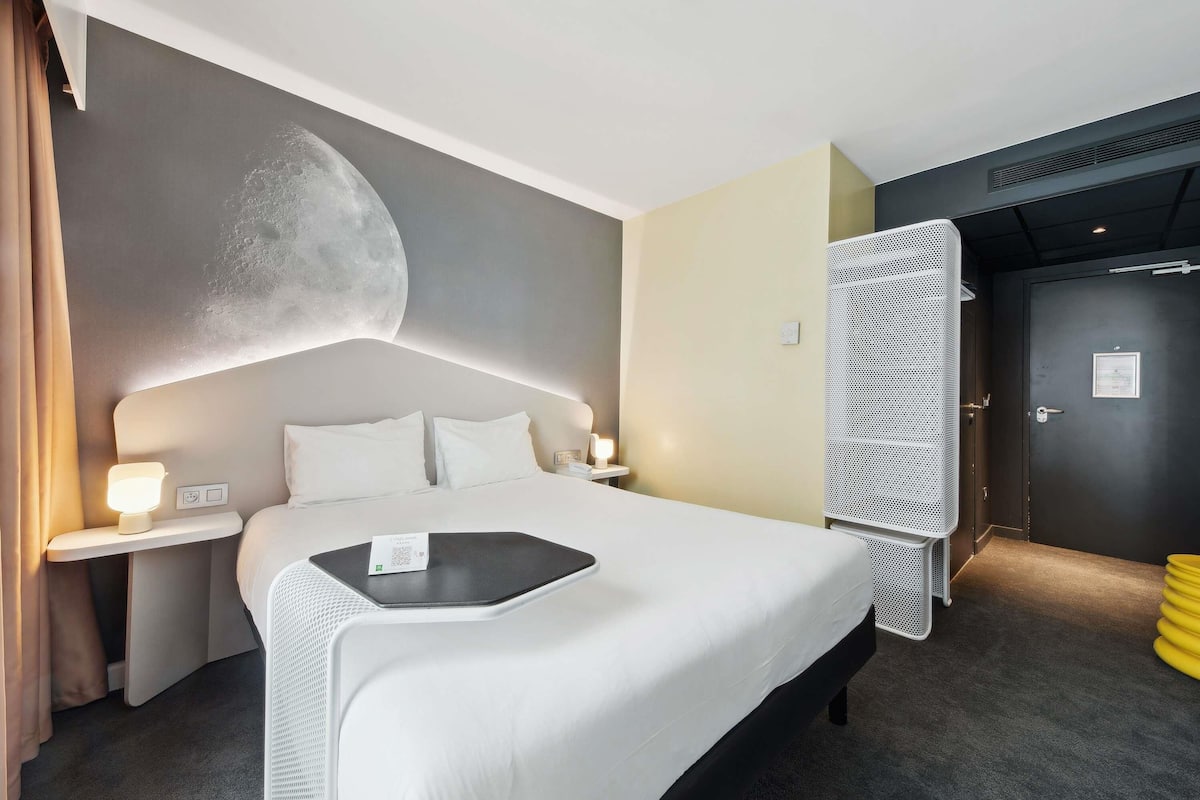 A hotel room with a bed, bedside tables, a wall-mounted moon design, a door with a metal mesh, and a dark-colored wall.