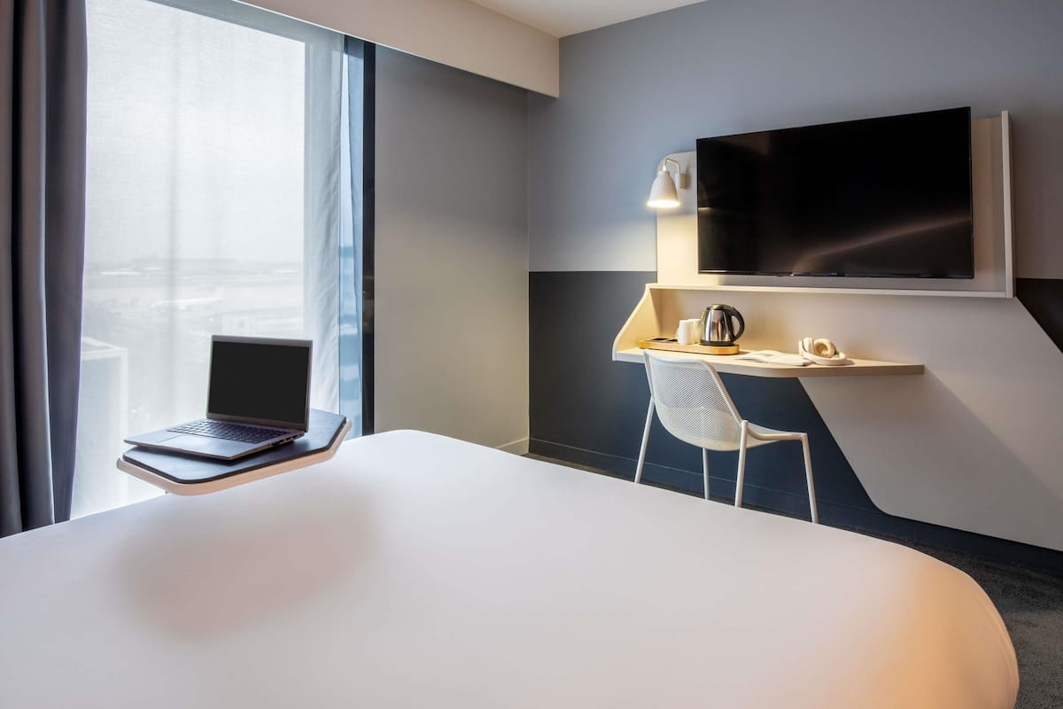Une chambre d’hôtel moderne dotée d’un grand lit, d’un bureau avec un ordinateur portable, d’une télévision à écran plat et d’une fenêtre avec des rideaux.
