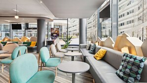 A modern lounge area with sofas, chairs, and tables, large windows offering views of the city, and a flat-screen TV mounted on the wall. - Ibis Styles Paris Charles de Gaulle Airport (Tremblay-en-France)
