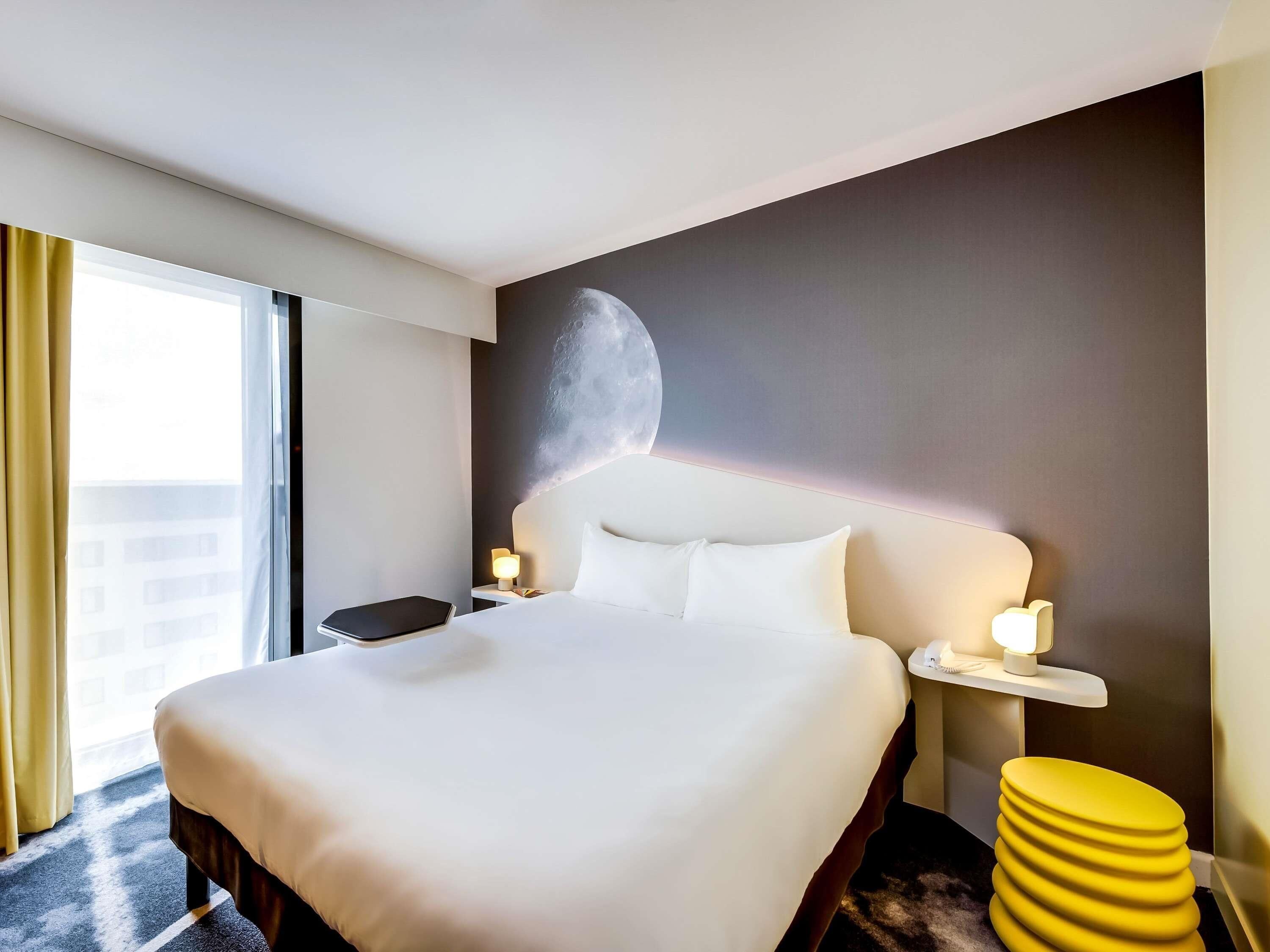 A hotel room with a large bed, a nightstand, a lamp, a yellow stool, and a wall with a moon mural.