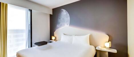 A hotel room with a large bed, a nightstand, a lamp, a yellow stool, and a wall with a moon mural.