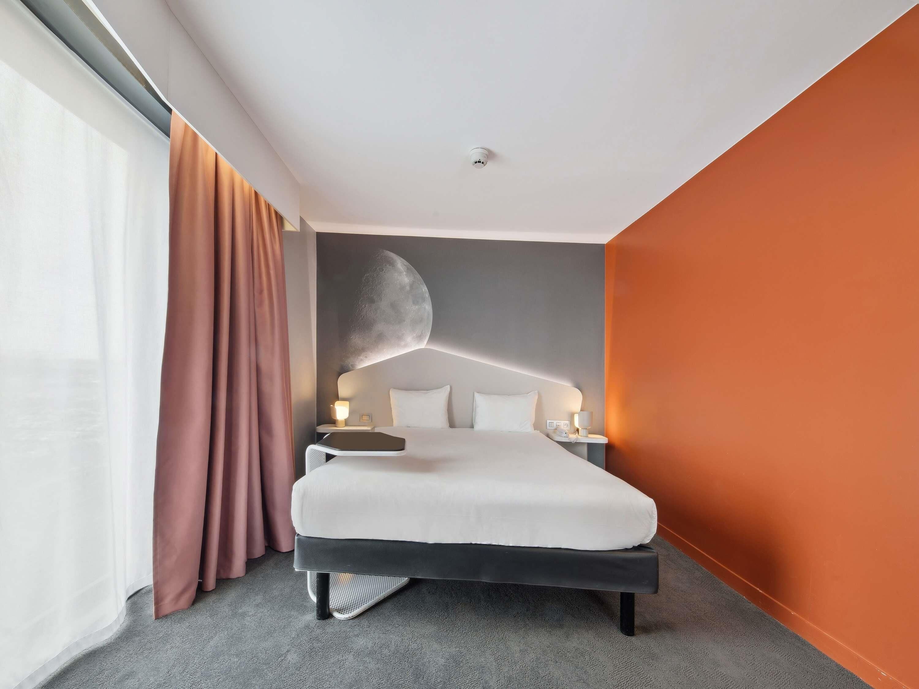 A modern hotel room with a large bed, a moon mural on the wall, a side table with lamps, and a large window with curtains.