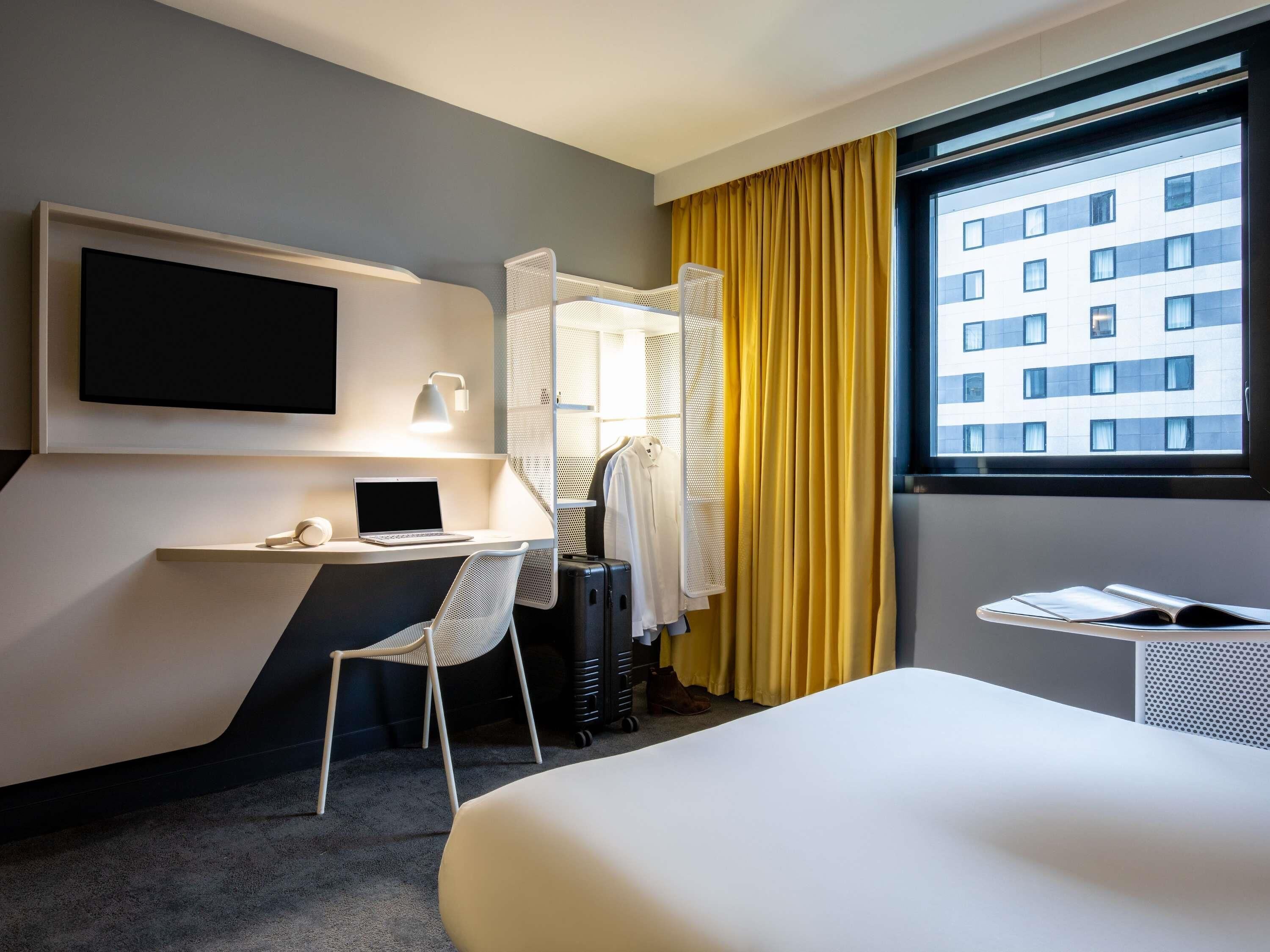 Habitación de hotel con una cama, un escritorio con una computadora portátil, una silla, un televisor y una ventana con cortinas.