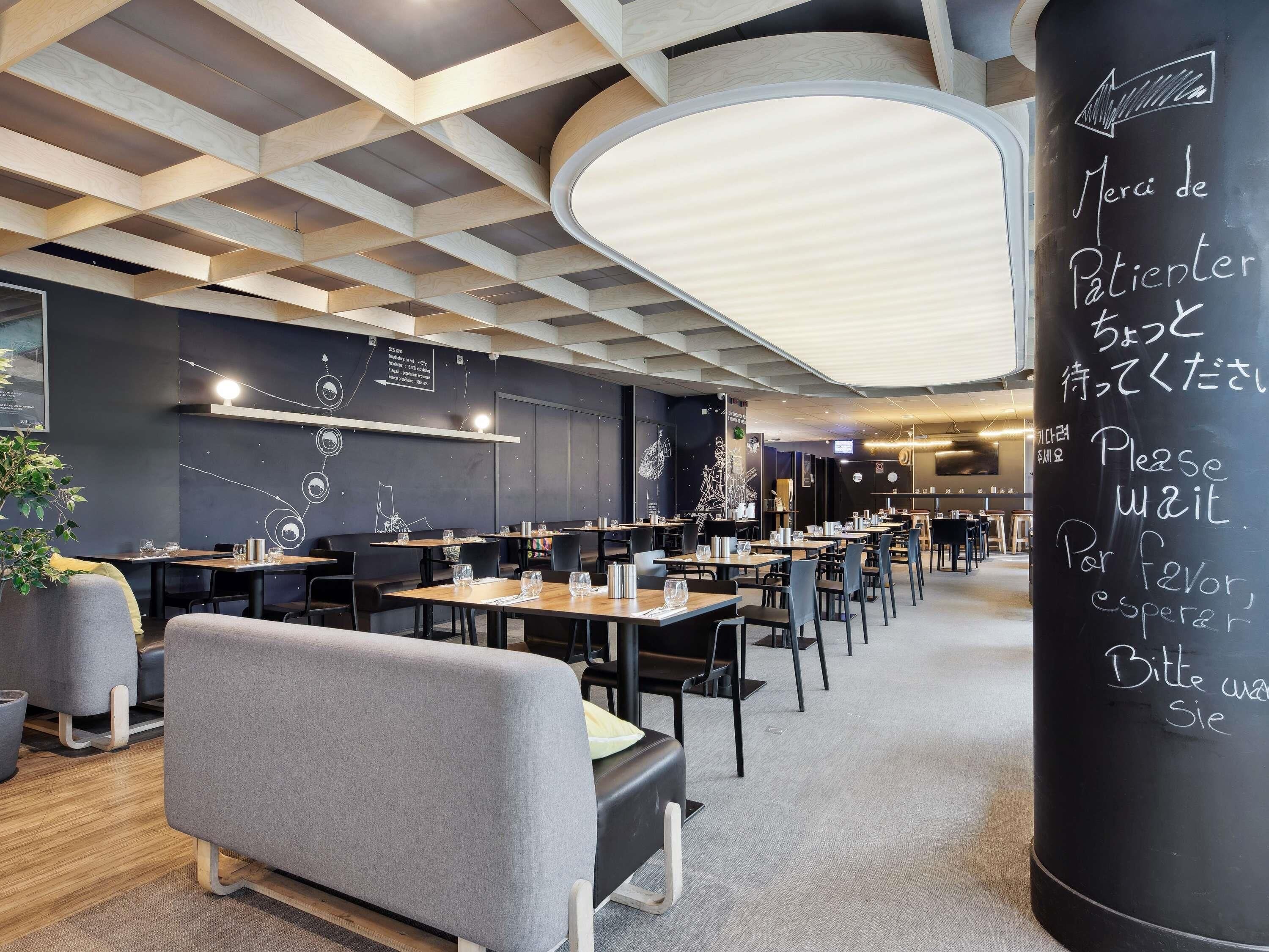 Un restaurant moderne, avec un intérieur caractérisé par un plafond au design géométrique, un tableau noir affichant des instructions multilingues pour la liste d’attente, ainsi que des tables et des chaises disposées de manière ordonnée.