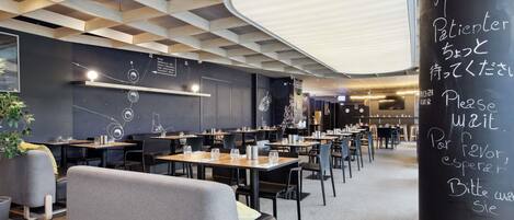 Un restaurant moderne, avec un intérieur caractérisé par un plafond au design géométrique, un tableau noir affichant des instructions multilingues pour la liste d’attente, ainsi que des tables et des chaises disposées de manière ordonnée.