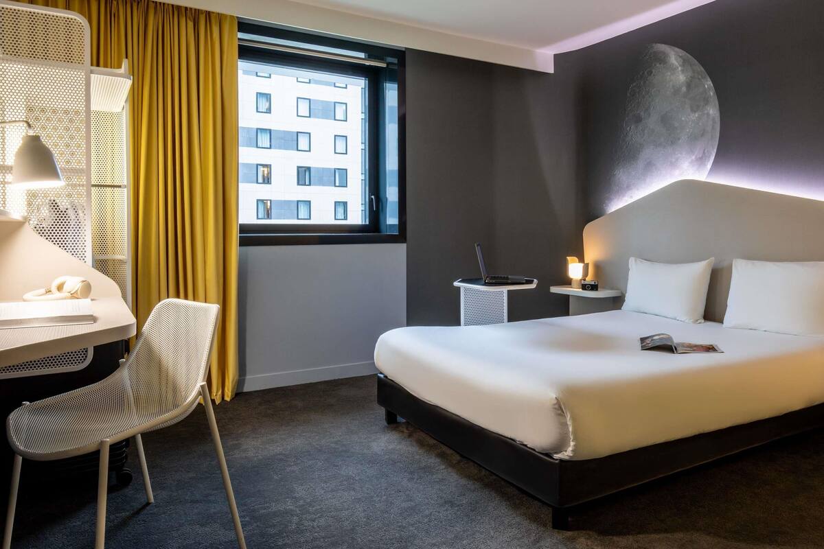 Une chambre d’hôtel avec un lit, un bureau, une chaise et une vue sur un immeuble par la fenêtre.