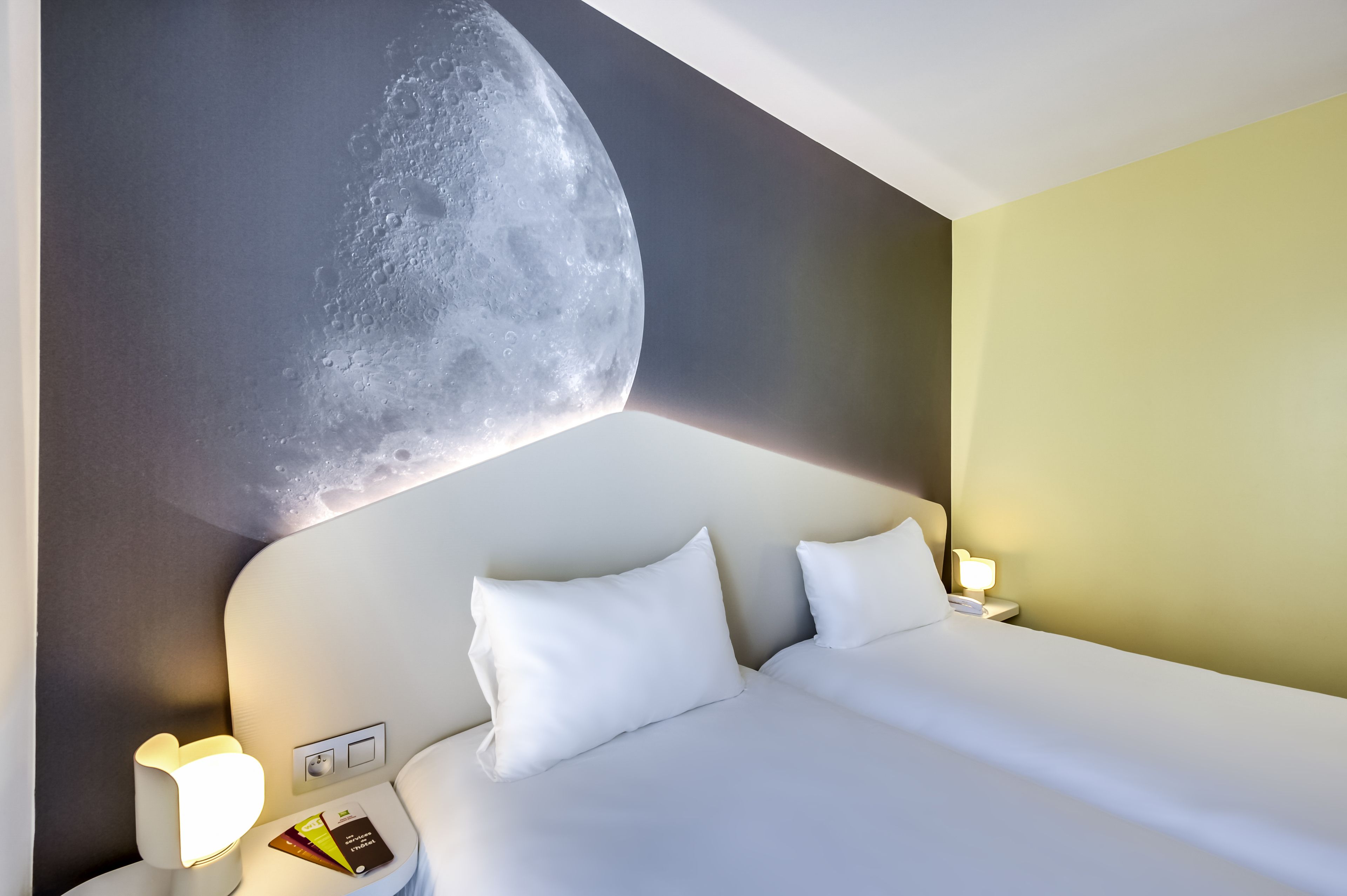 A hotel room with a bed, white pillows, a nightstand with a lamp, and a wall mural of the moon.