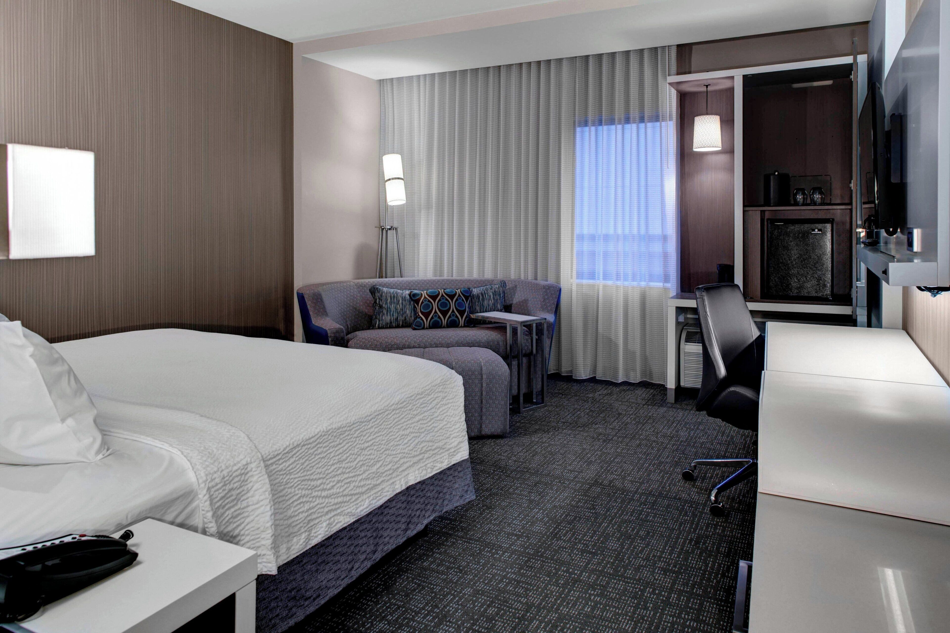 A hotel room with a bed, a sofa, a desk, a chair, a TV, and a minibar.