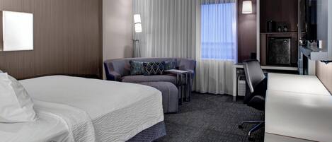 A hotel room with a bed, a sofa, a desk, a chair, a TV, and a minibar.