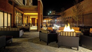 An outdoor patio area with wicker furniture, a central fire feature, and a brick building in the background. - Courtyard Carrollton (Carrollton)