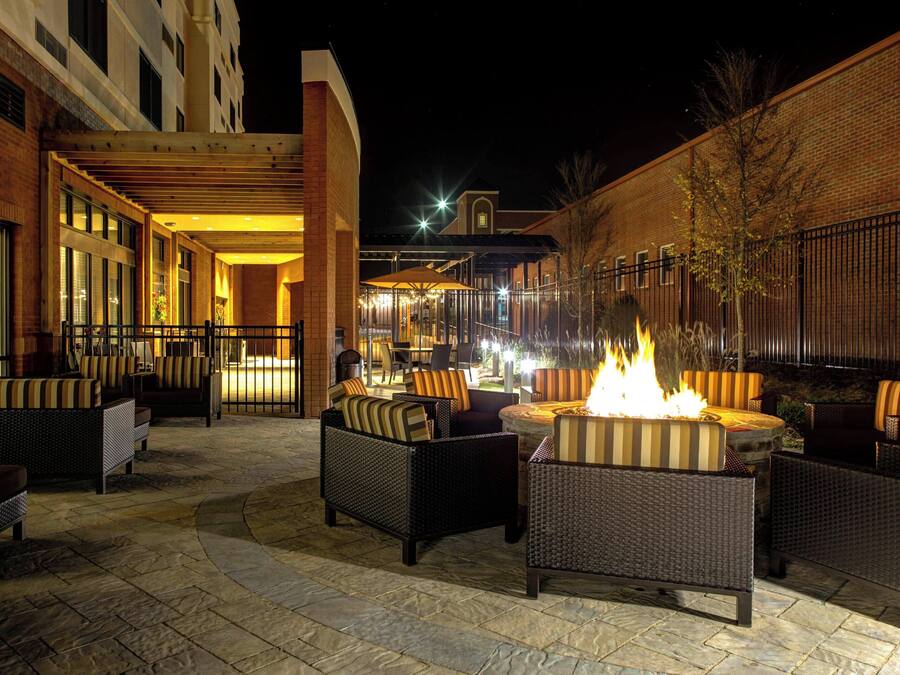 An outdoor patio area with wicker furniture, a central fire feature, and a brick building in the background.