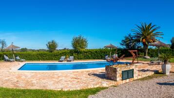 Piscina con sillas de descanso y sombrillas de paja, rodeada de baldosas de piedra y vegetación.